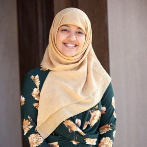 Olive skinned woman wearing a beige hijab and dark green dress with gold accents.